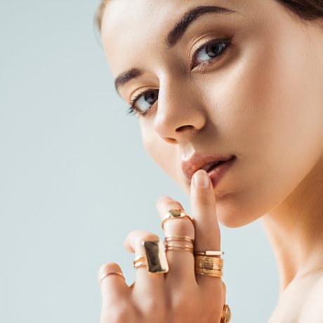young woman with shiny makeup and golden rings touching lips isolated on grey